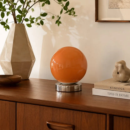 Lampe de bureau Ignis à globe orange posée sur un buffet en noyer parmi des objets céramique et un vase minéral